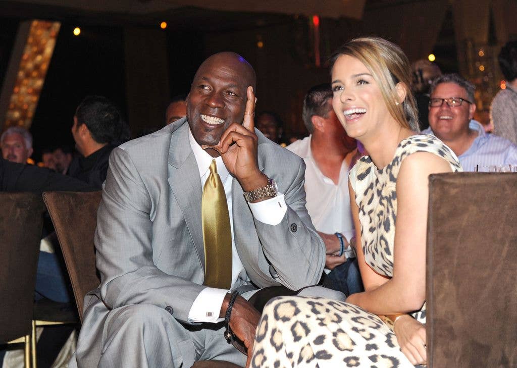 Michael Jordan in a grey suit and yellow tie sat down smiling, with his fiancee Yvette Prieto next to him laughing, wearing a long lepoard print dress 