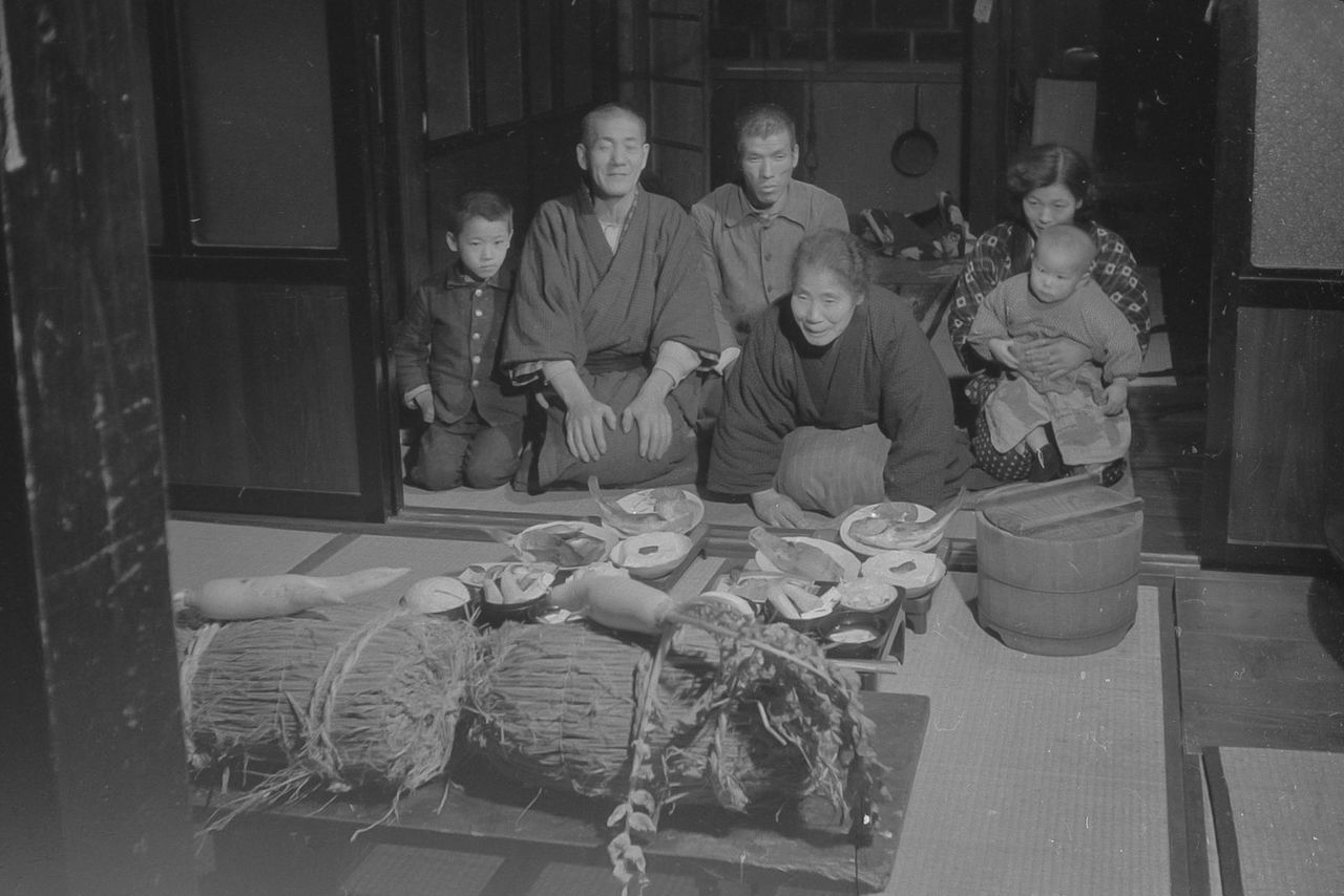 A family offers food and drink to the tanokami, represented here by bales of seed rice and a forked daikon radish. (© Haga Hideo)