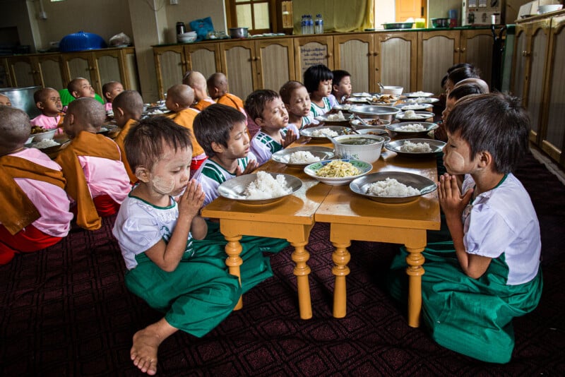 Young children with traditional face paint sit cross-legged in groups around low tables, hands clasped in prayer, with plates of rice and food in front of them, inside a communal dining hall.