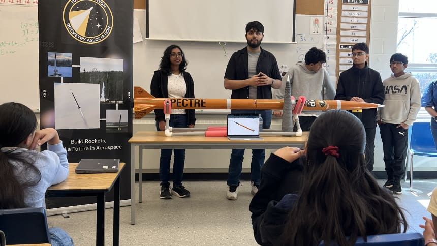Five students stand in front of a classroom with an orange rocket displayed on the table in front of them.