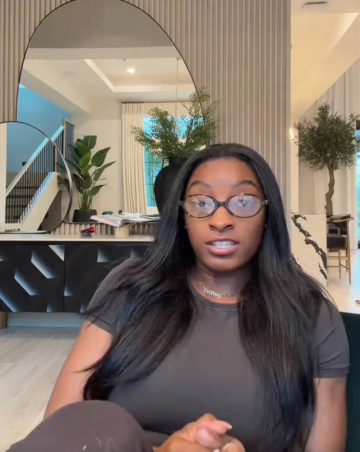 A person in a casual outfit and glasses speaks to the camera in a modern living room setting