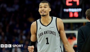 Victor Wembanyama holds up his fist and is wearing a grey vest
