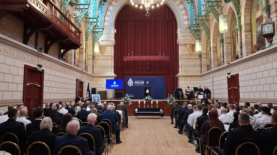 Another view of the civil service as dozens of people sit in rows in a hall, with a high-ceiling, with an aisle down the middle. The people have their backs turned, facing the stage.