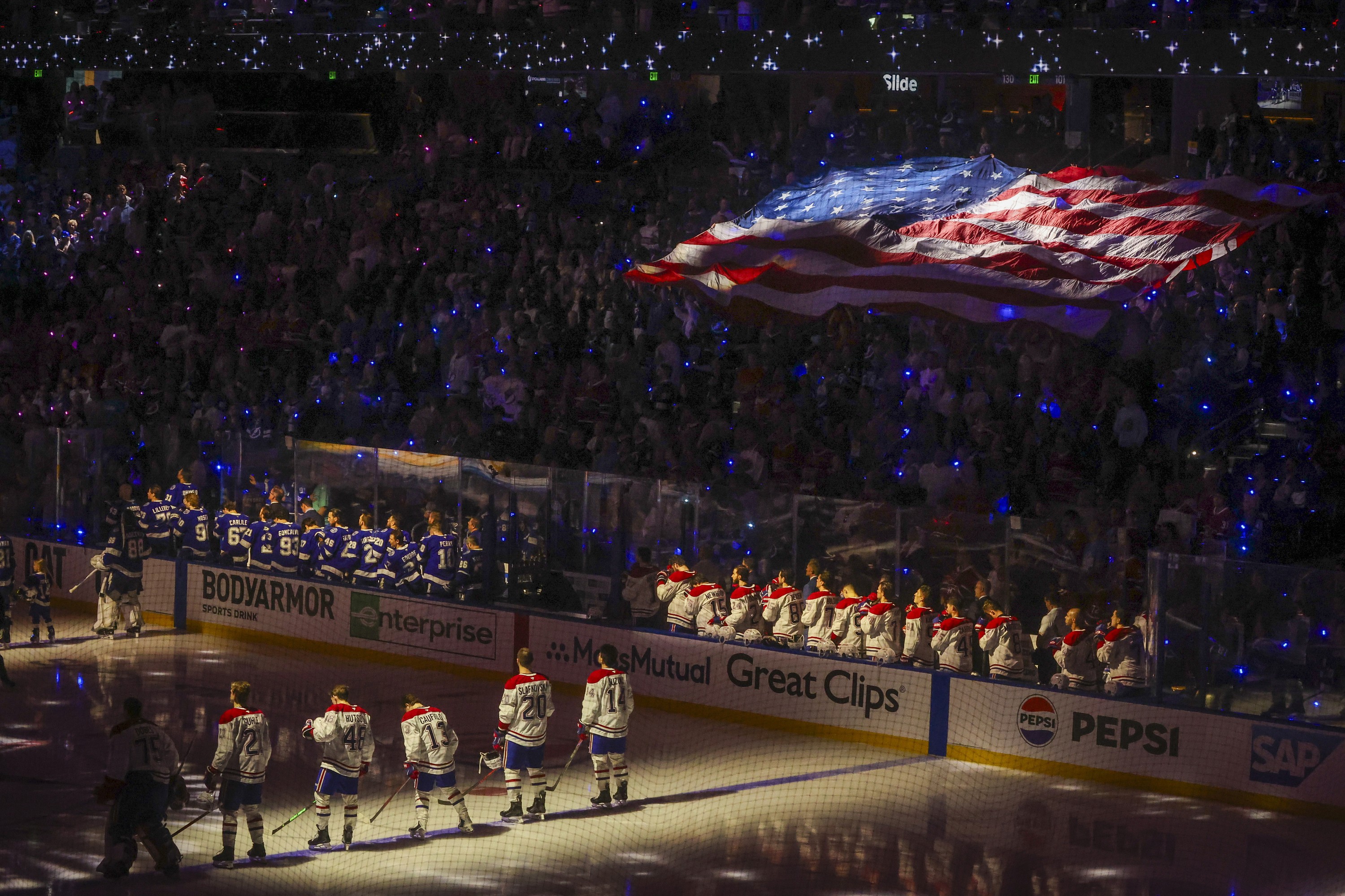 Tampa Bay Lightning and Montreal Canadiens players on the bench and on the blue lines as a large American flag is seen in the crowd and several small blue lights are lit in the stands.