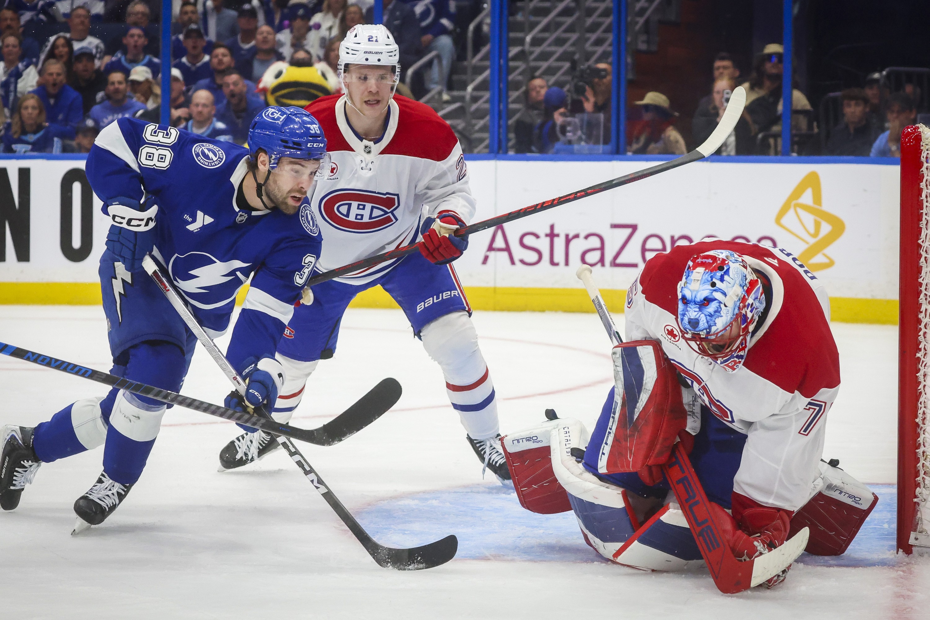 A Canadiens goaltender bends over to block a shot in the crease as a Lightning player skates toward him, and a Canadiens player behind him.