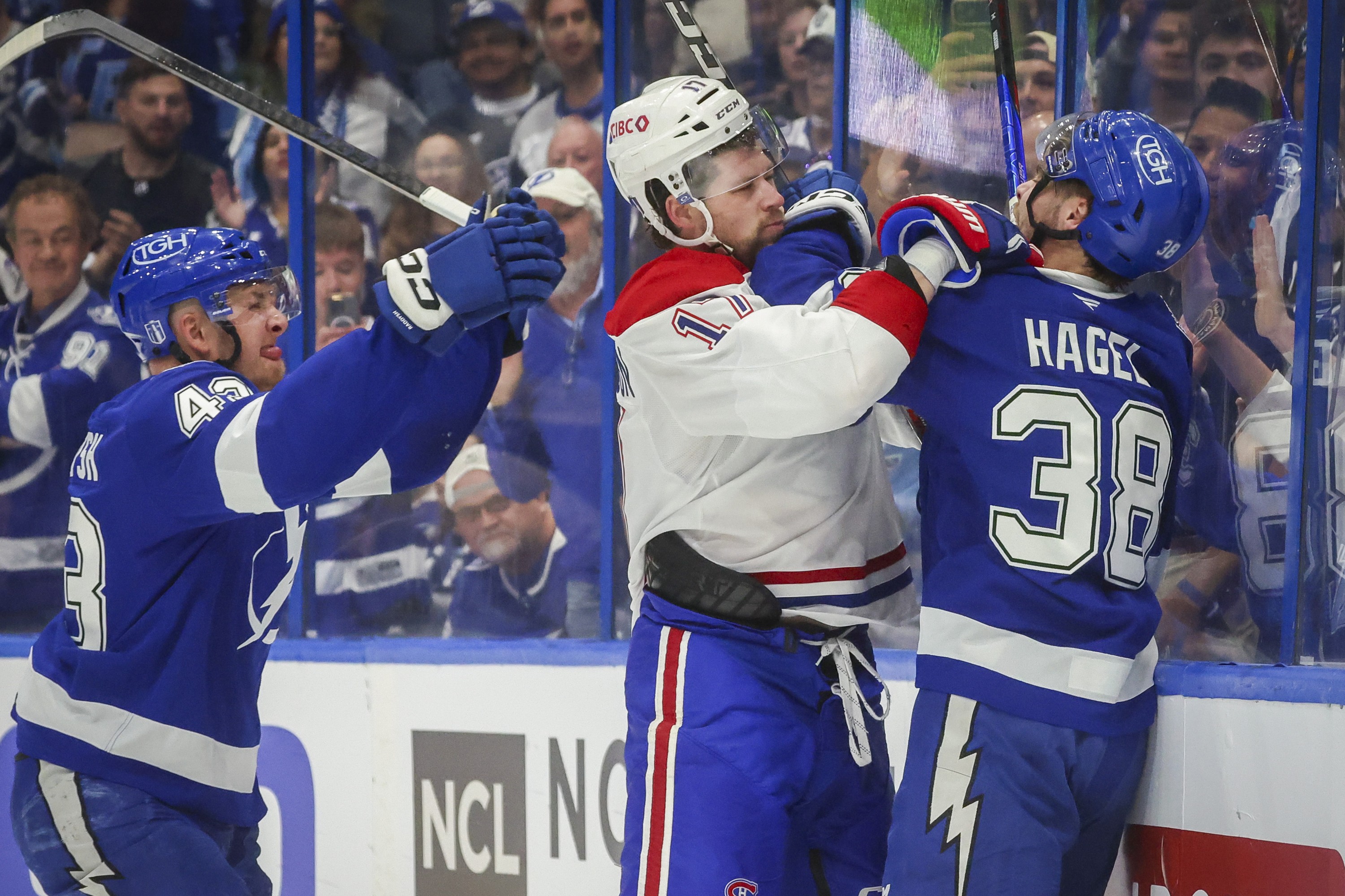 A Canadiens and Lightning player grab each other's shoulders along the boards as another Lightning player approaches with his stick in the air