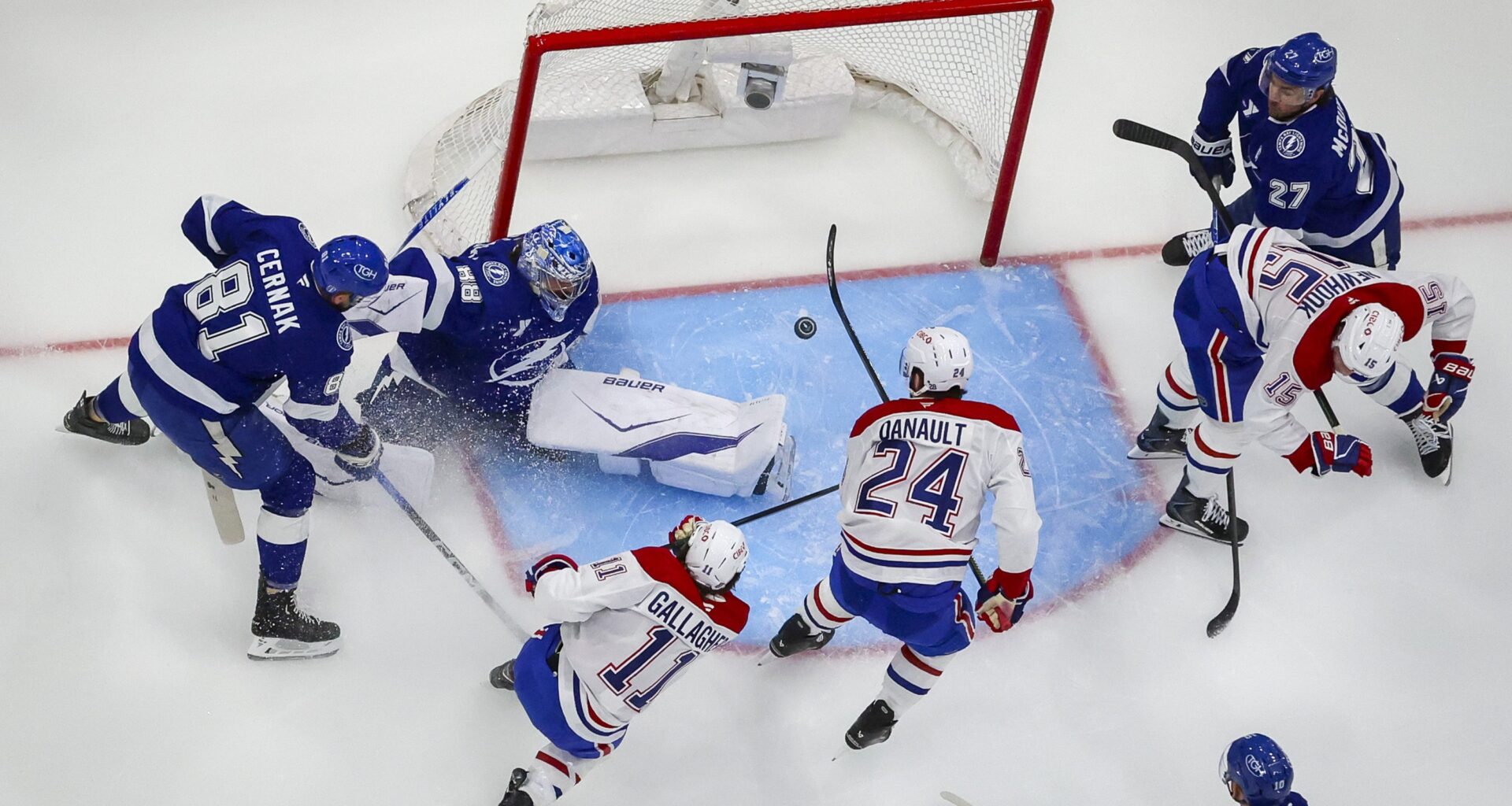 An overhead photo shows the puck mid-air in front of a mostly empty net as Brendan Gallagher follows through on a shot and several other players are around the crease.