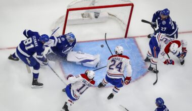An overhead photo shows the puck mid-air in front of a mostly empty net as Brendan Gallagher follows through on a shot and several other players are around the crease.