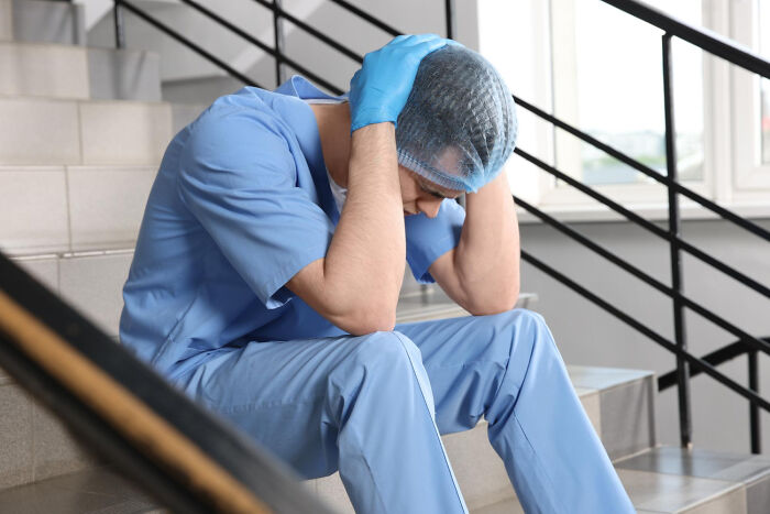 Healthcare worker in blue scrubs sitting on stairs holding head, illustrating hidden truths about hospitals and their harsh realities.