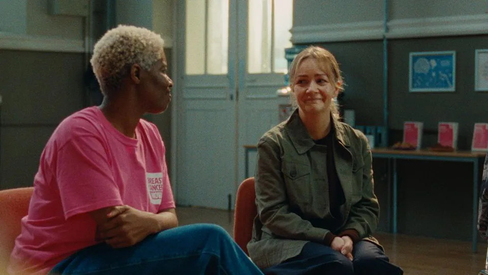 Two women sitting in a room, smiling at each other. One of them is wearing a pink T-shirt with the logo of Breast Cancer Now.