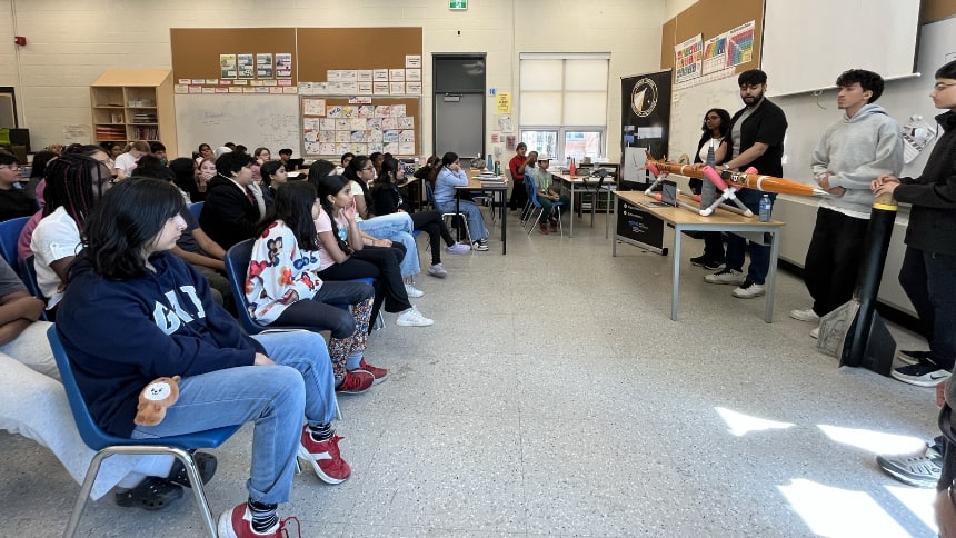 Students sitting in chairs look at a group of teenagers at the front of a classroom with an orange rocket in front of them. 