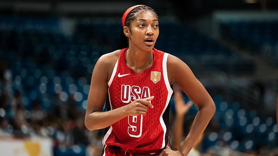 Angel Reese stands on an indoor court wearing a red USA basketball jersey with the number nine on the front and a red headband. One hand rests near the waist while the other arm is raised slightly, pointing forward. Blue arena seats and blurred figures are visible in the background under bright lighting