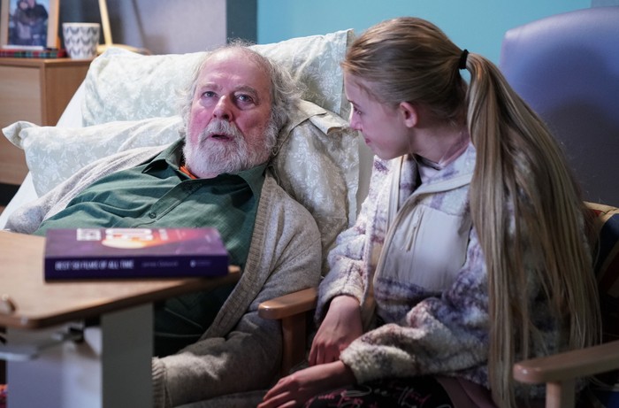 Lexi sits beside Nigel, who is lay in bed, in a scene from EastEnders