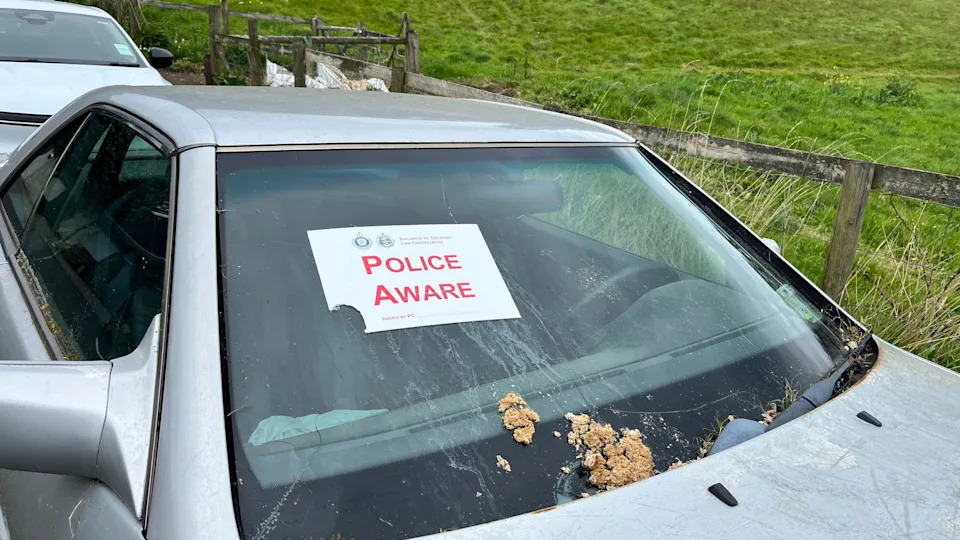 A silver car parked near a grassy area with a wooden fence, displaying a “Police Aware” notice from Bailiwick of Guernsey Law Enforcement on its front windshield