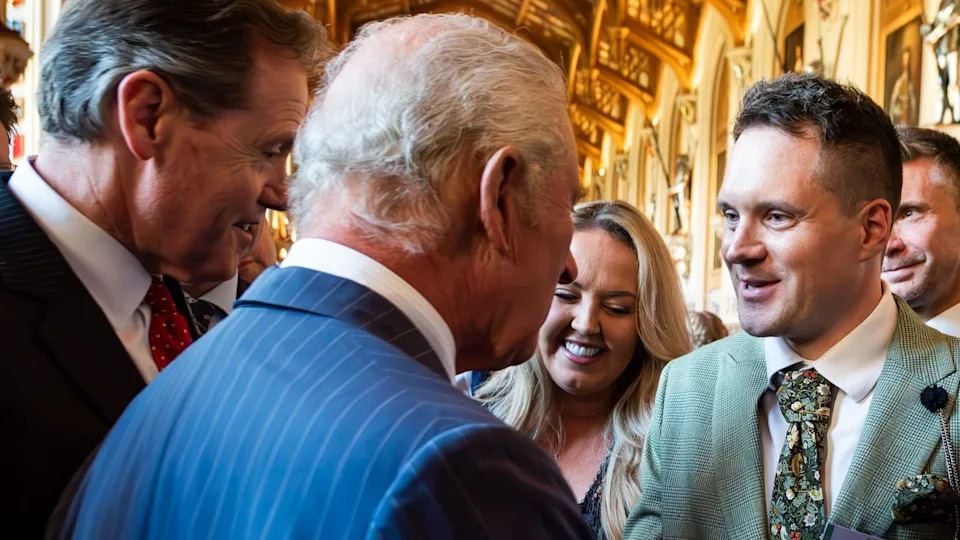 A close up of the back of King Charles III's head as he talks to Joseph Holman. The king is wearing a blue, pinstriped suit. Holman is wearing a mint-green suit, white shirt and floral tie.