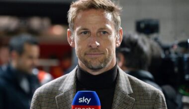 Button in high-neck dark top and beige blazer in the F1 paddock holding a Sky Sports microphone