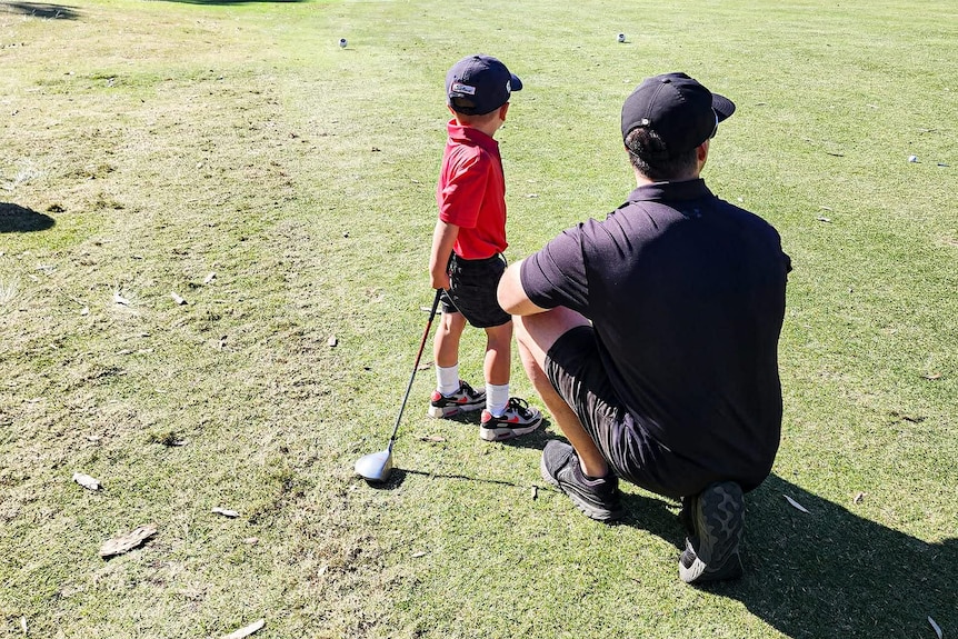 A young boy with his golf club stand beside a man who is crouched next to him