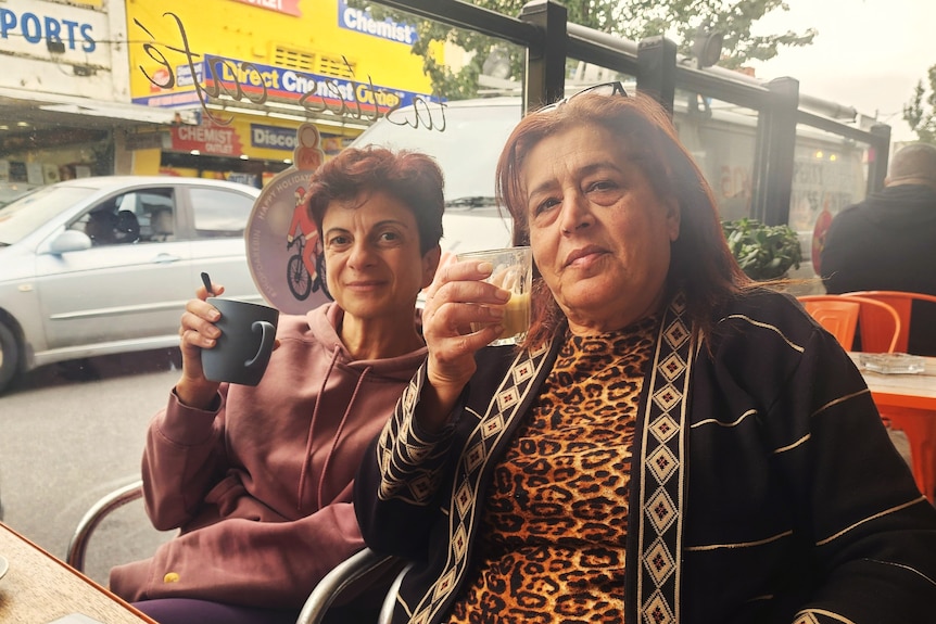 Two women sit at a cafe on a busy street, drinking coffee. 
