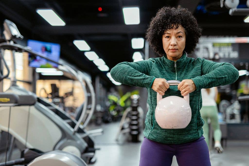 A Middle-aged, Woman Working Out With a Kettlebell in the Gym. She Holds the Weight Confidently, Demonstrating Strength and Focus During Her Exercise. Her Workout Clothes Are Practical and Supportive, and Her Facial Expression Shows a Sense of Accomplishment.