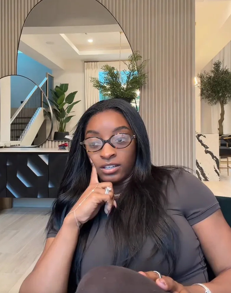 A person with glasses and long hair sits in a stylish modern room, appearing engaged or thoughtful
