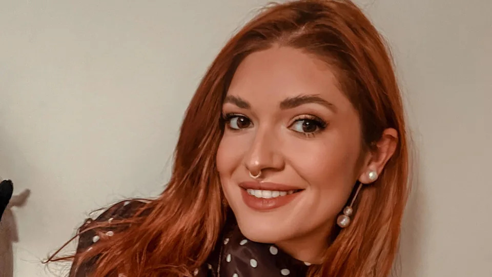 A selfie of a young woman with long red hair. She is wearing a brown silk dress with white polka dots and has tattoos on both arms.