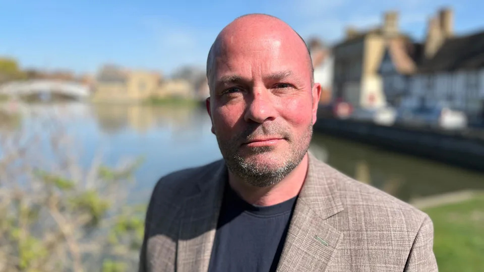 Phillip Malley looking into camera in front of the waterfront in Godmanchester. He has stubble and is wearing a brown suit and black t-shirt.