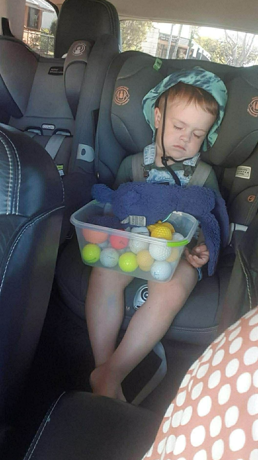 A young boy sitting in a child's car seat holding a tupperware container full of golf balls