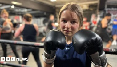 A 19 year-old woman is standing next to a boxing ring. She is wearing black boxing gloves which she is holding up close to her face. She has light brown hair tied back and is wearing a navy T-shirt. Other poeple are training in the ring behind her.