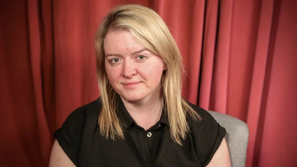 A woman with blonde hair in a black top sits on a chair in front of a red curtain