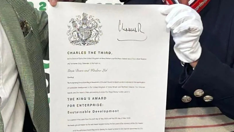A close up of the certificate from King Charles III, complete with signature and his coat of arms. It is being partly held up by Susan Lousada, the Lord-Lieutenant of Bedfordshire, who is wearing white gloves.