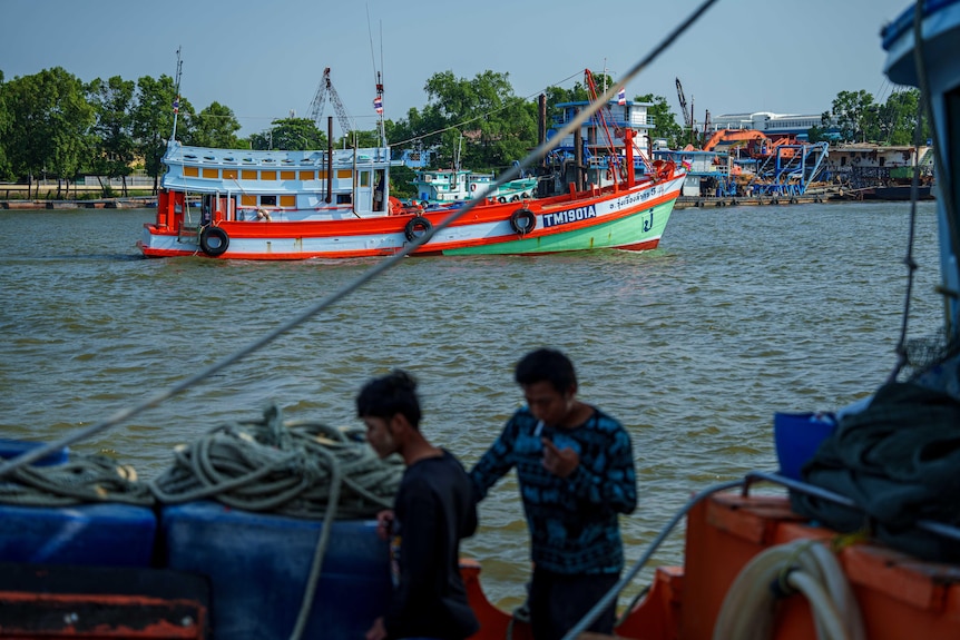 Two people, one smoking a cigarette, walking along a dock laden fishing paraphenalia, while a boat sails in the background