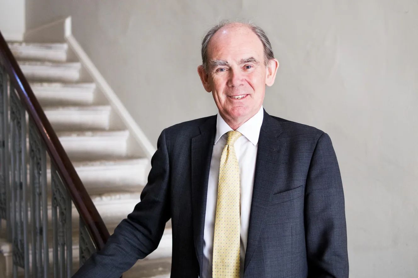 Paddy Grafton Green smiling in a dark suit with a yellow tie, standing next to a staircase.