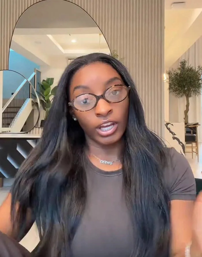 A person is speaking in a modern, stylish interior. They have long hair, glasses, and are wearing a simple top