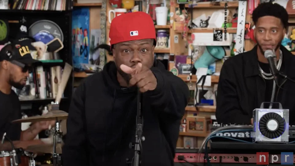 Kevin Hart and two musicians performing at an NPR Tiny Desk Concert. The central figure wears a red cap and points at the camera, with instruments around.