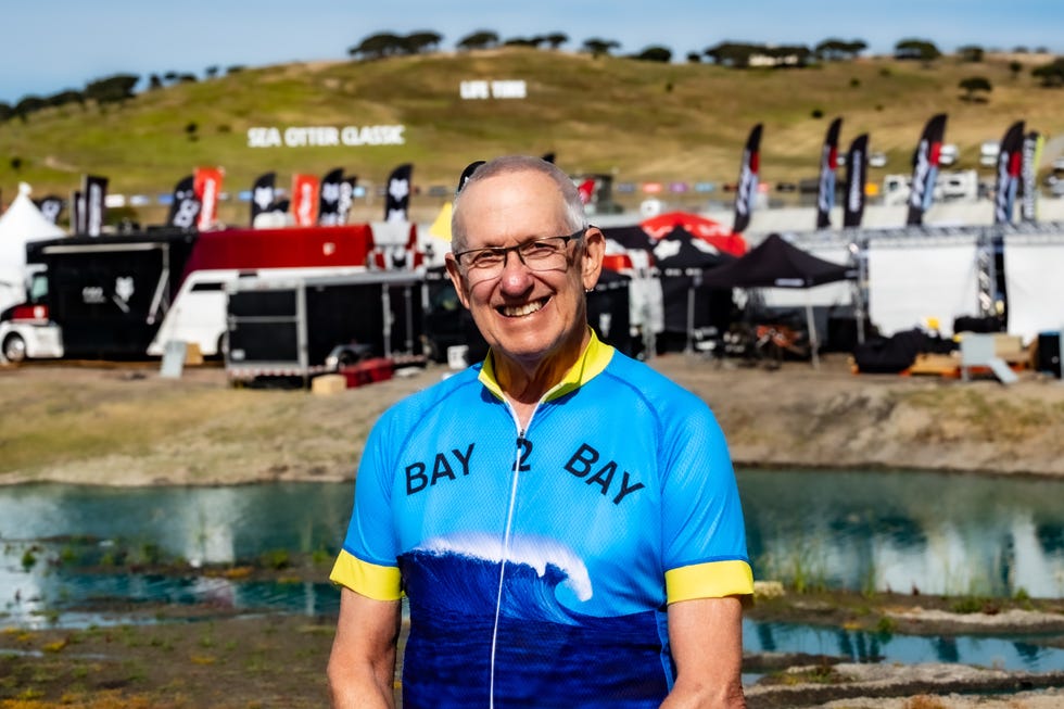 Cyclist in a vibrant blue and yellow jersey with a wave design stands near an event setup. Cyclist in a vibrant blue and yellow jersey with a wave design stands near an event setup.