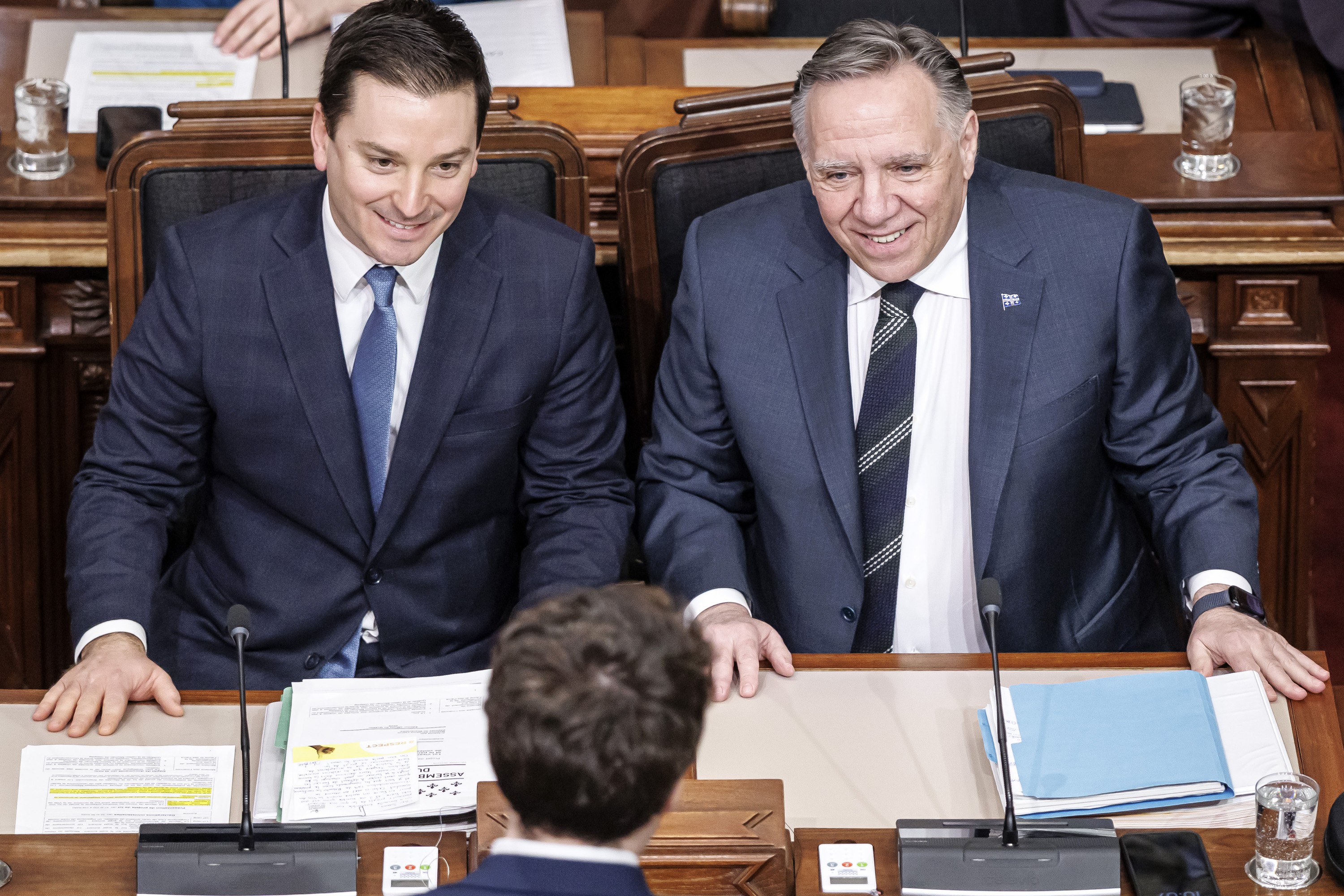 Simon Jolin-Barrette and François Legault smile while sitting next to each other at their desks in the National Assembly's red room.