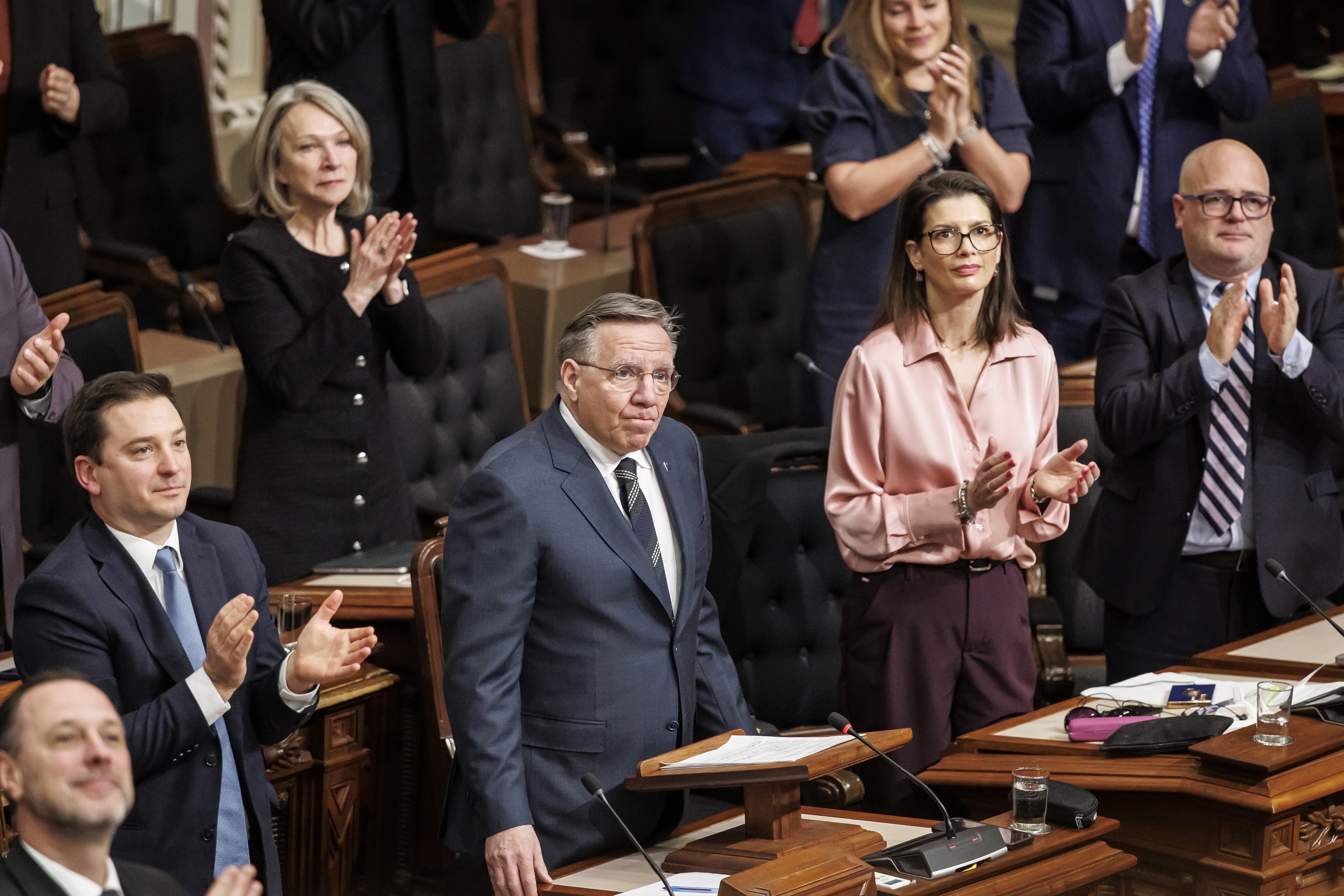 François Legault looks up while standing at his desk at the National Assembly's red room while MNAs around him applaud.