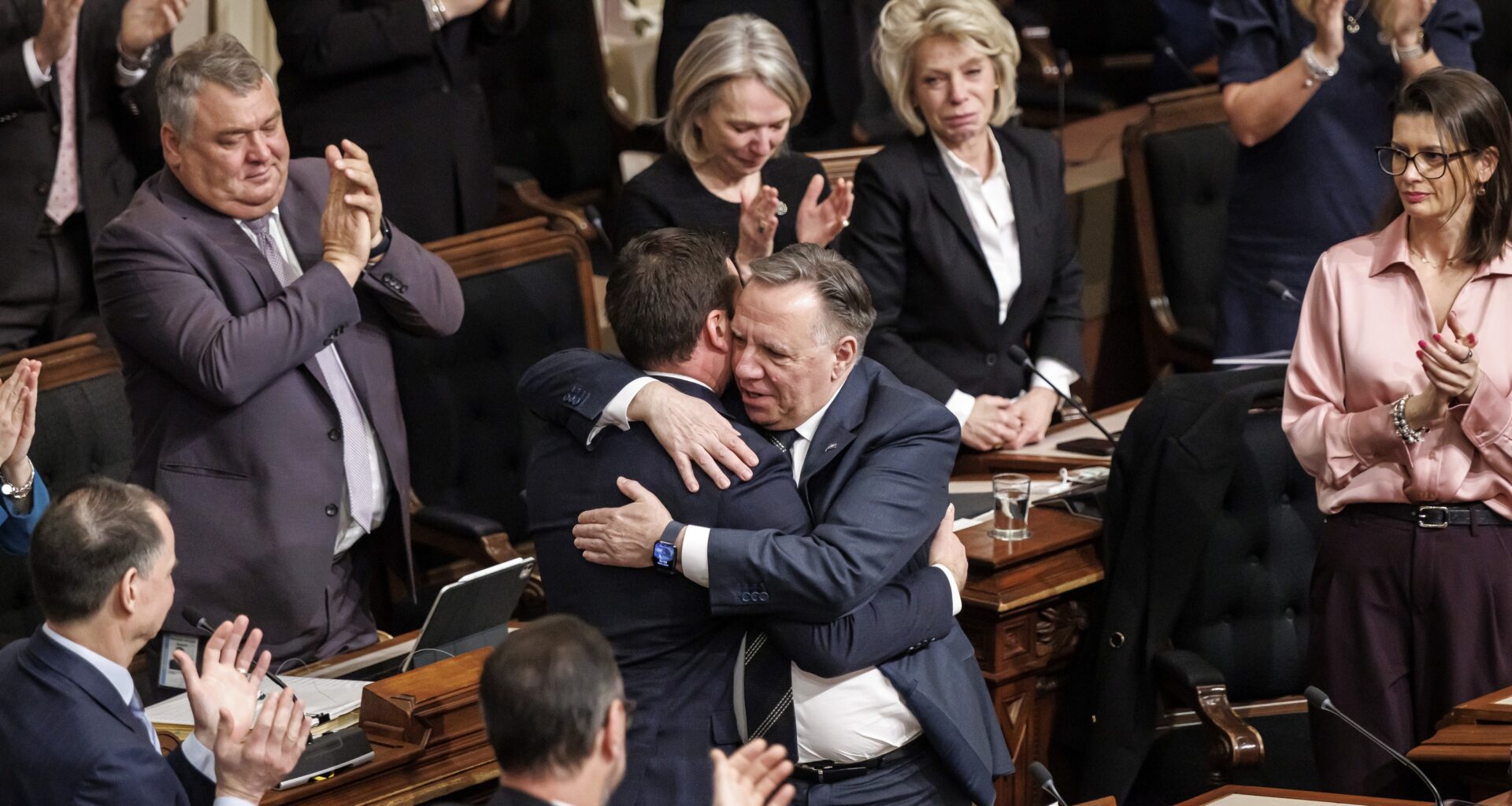 François Legault hugs Simon Jolin-Barrette as MNAs at their desks around them applaud.