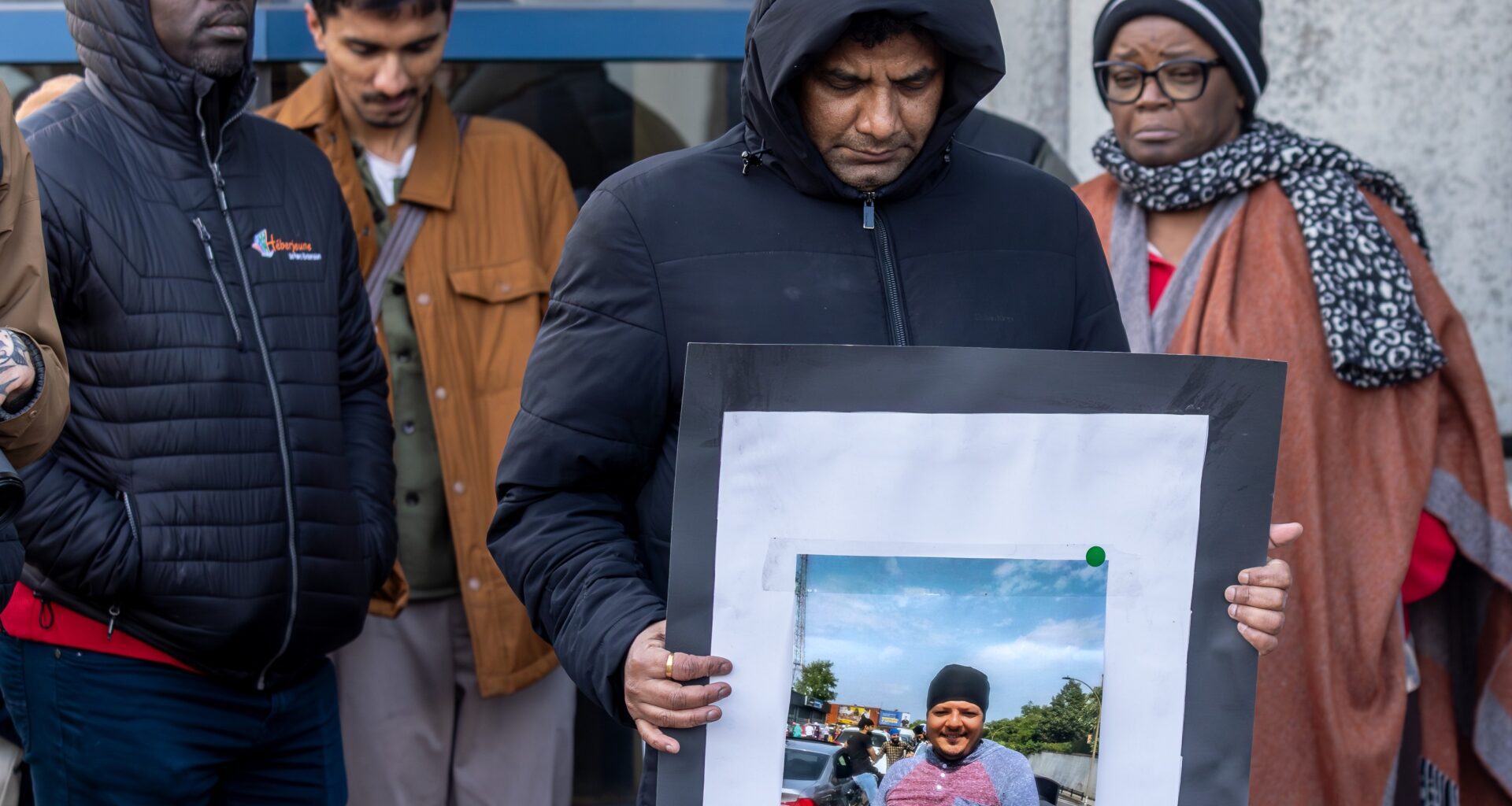 Four people stand with heads bowed. One man is holding a photo of an unhoused man.