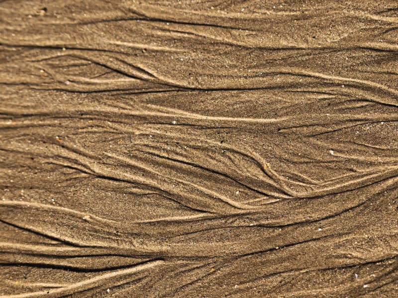 Close-up of wet sand with natural, wavy patterns and grooves formed by water flow, creating an abstract, textured design in light and dark brown tones.