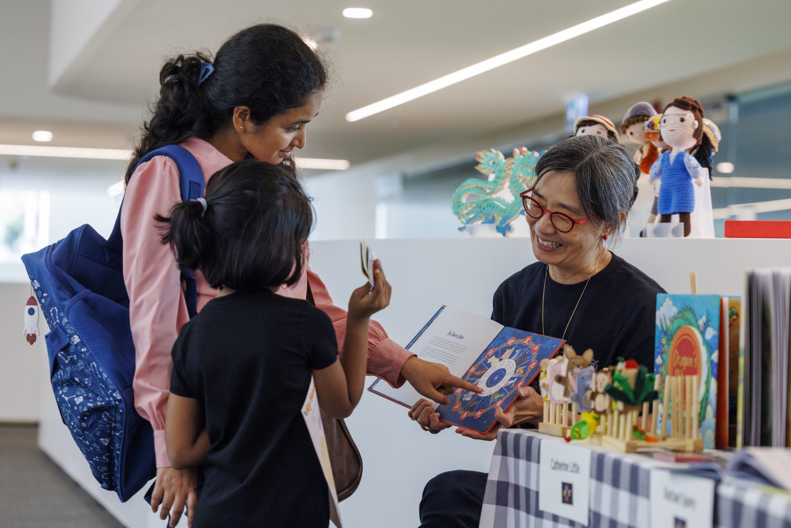 An older adult seated at a display table shares an illustrated book with an adult and a child inside a public library, with more books and small decorations arranged on the table.