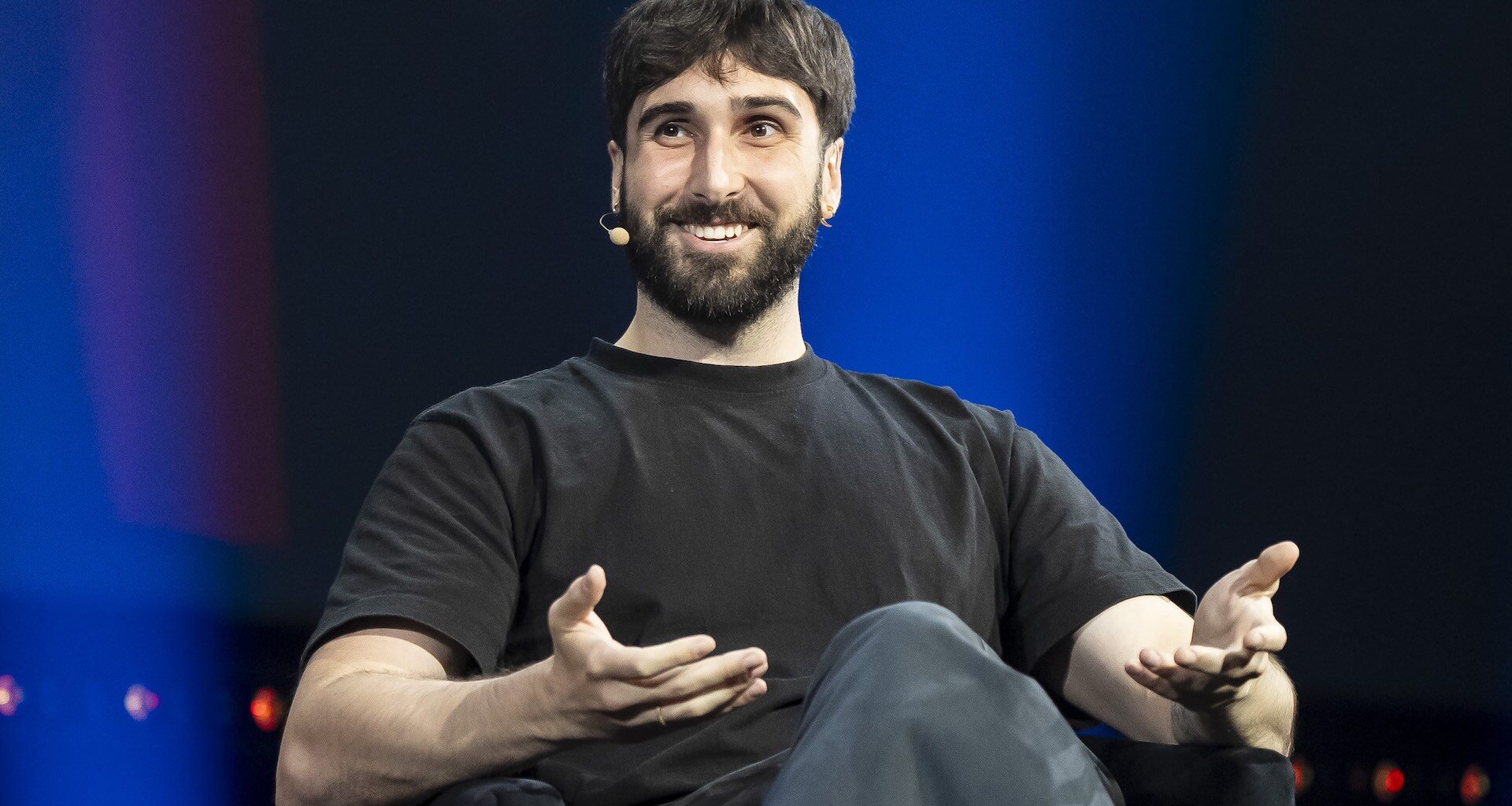 A man wearing a headset microphone sits on a stage. He is speaking and gesturing with his hands. The background is dark blue.
