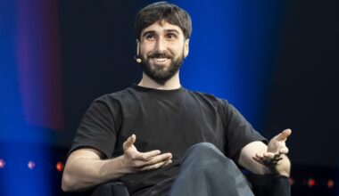 A man wearing a headset microphone sits on a stage. He is speaking and gesturing with his hands. The background is dark blue.