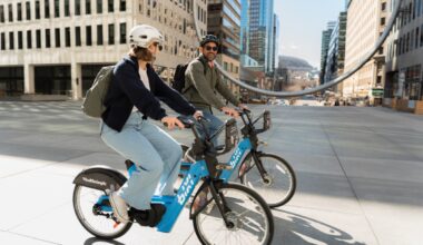Two people wearing helmets ride blue shared bikes through a sunny city plaza with tall buildings in the background, backpacks on.