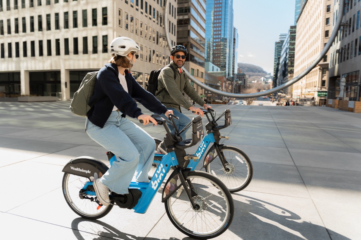 Two people wearing helmets ride blue shared bikes through a sunny city plaza with tall buildings in the background, backpacks on.
