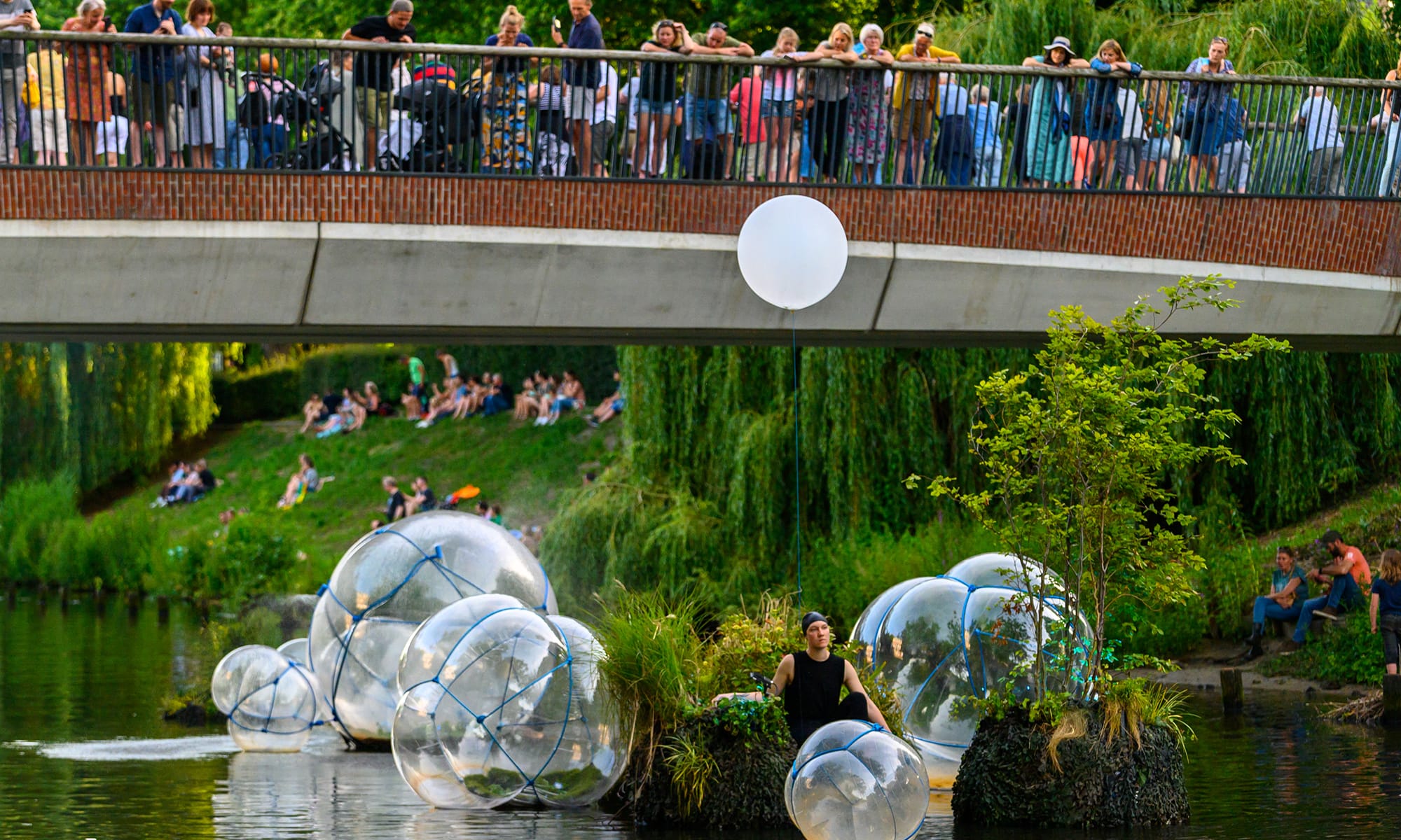 Photograph of a person at the center of a floating artwork consisting of large, translucent bubbles and grass covered mounds.