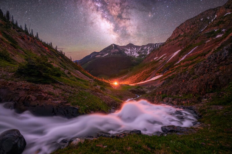 A bright Milky Way shines over a mountain valley with a glowing campfire in the distance, snowy peaks, and a stream flowing through rocky terrain in the foreground at night.