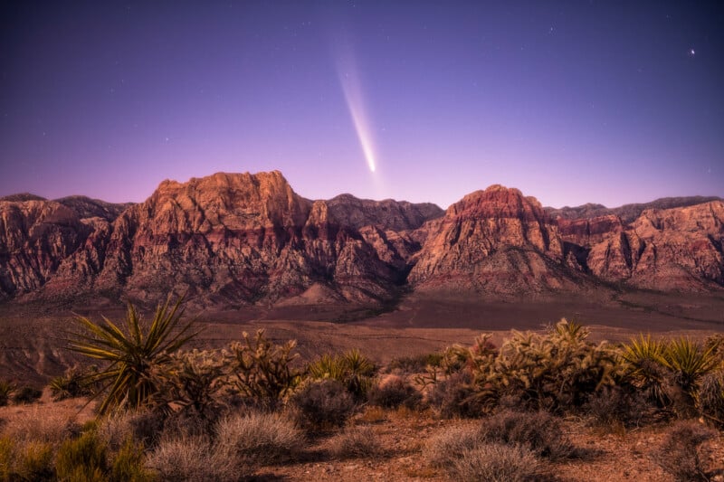A bright comet with a long tail streaks above rugged red rock mountains at dusk, with desert plants and cacti in the foreground under a clear, star-speckled sky.