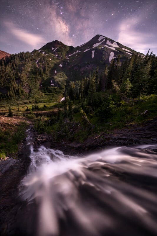 A mountain landscape with patches of snow, lush green trees, and a stream flowing over rocks in the foreground. The sky above shows stars and the Milky Way, adding a nighttime ambiance.