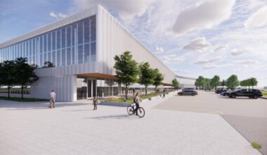 Modern commercial building with glass windows, trees, people walking and biking, and a parking lot under a partly cloudy sky.